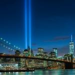 The Tribute in Light rises over Lower Manhattan behind the Brooklyn Bridge at night.
